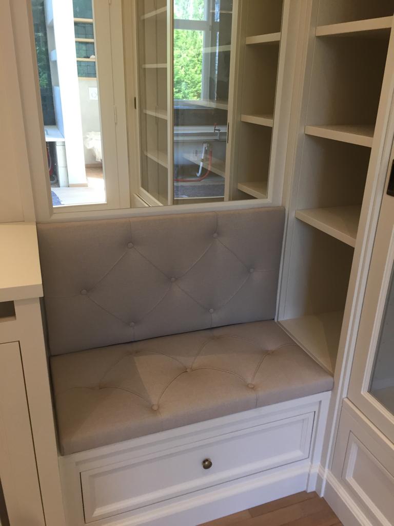 Banquette d'encastrement capitonnée en lin gris clair, intégrée dans une niche de bibliothèque blanche avec tiroir